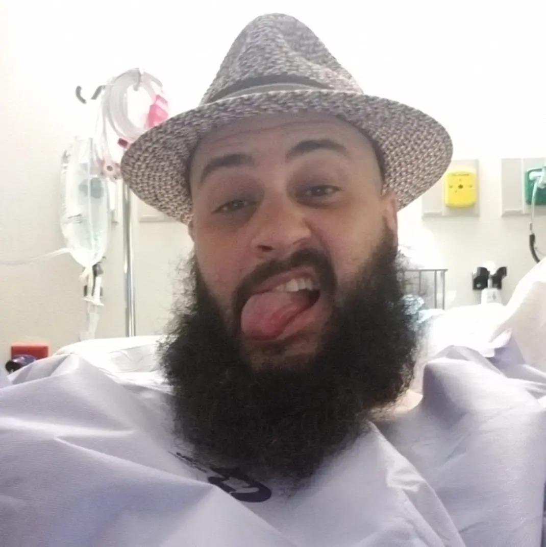 Jeremy Ryan in a hospital gown and woven hat, tongue out, IV bags behind him.
